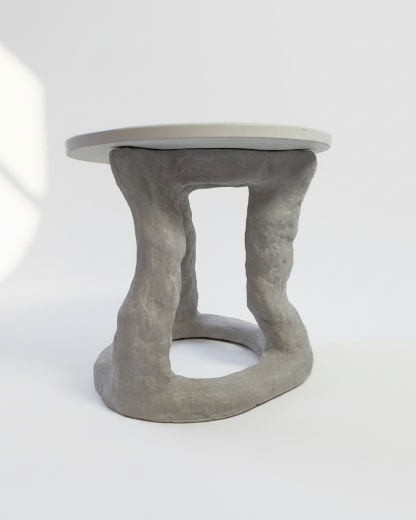 A sculptural side table with a round light gray top and a textured, organic-shaped gray base, shown on a clean white studio background with soft window shadow overlays.