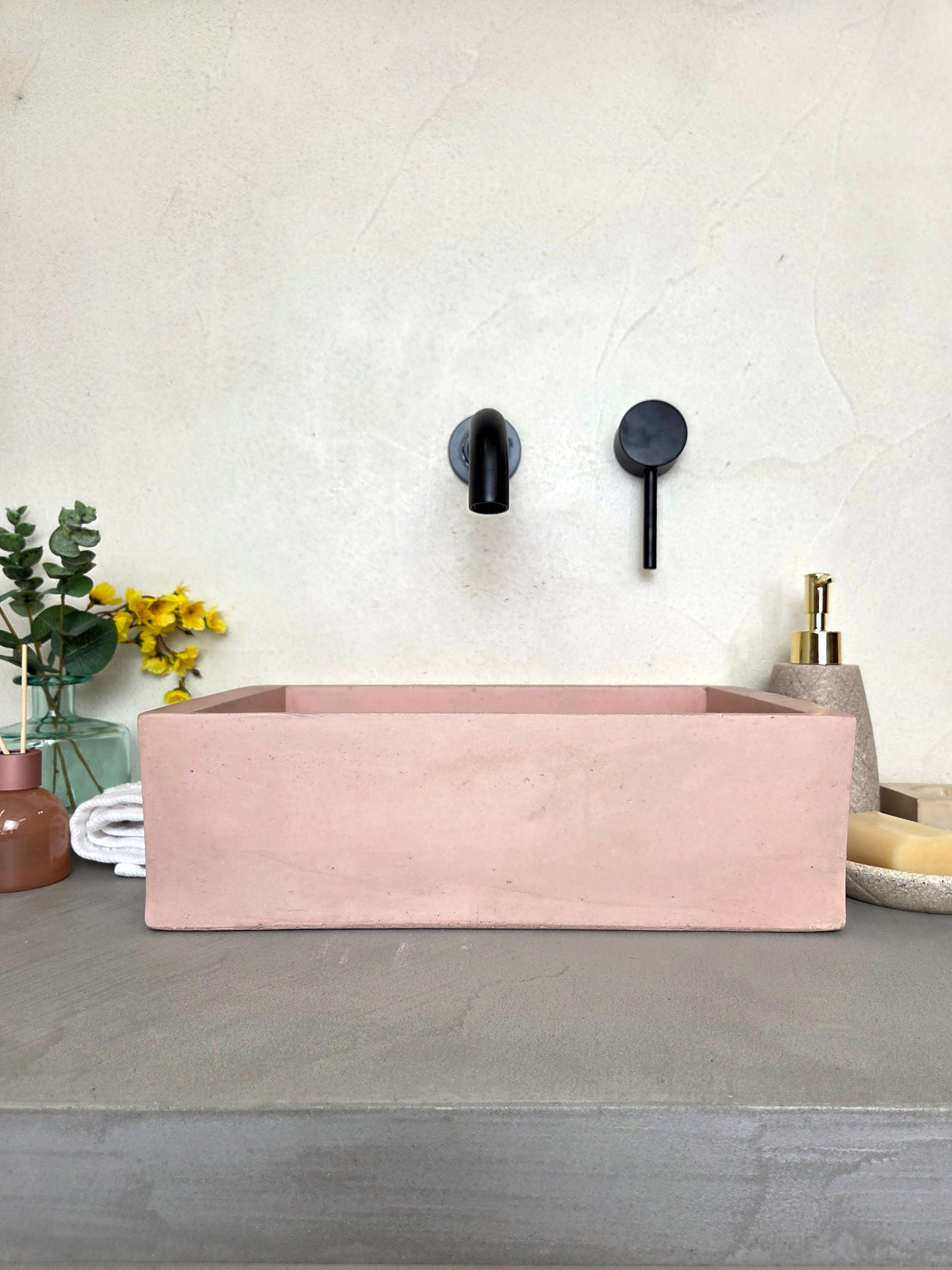 MINI rectangular concrete sink in pink, on a gray countertop with black wall-mounted fixtures.