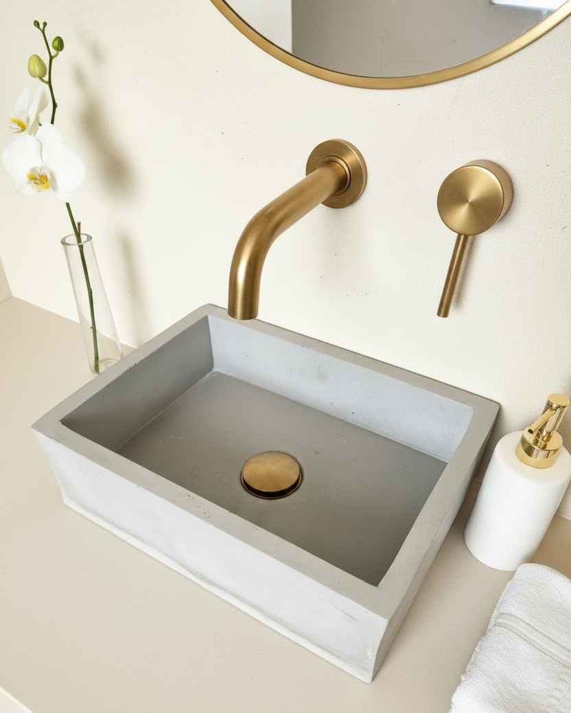 A modern rectangular concrete vessel sink with a matte gray interior and a brushed gold pop-up drain, mounted on a beige countertop with a matching gold wall-mounted faucet and lever handle.