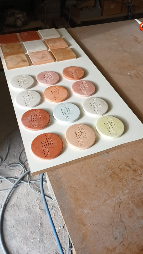 A white rectangular board displaying multiple circular and square concrete colour sample pieces in assorted earthy colors, photographed on a workshop table with cables and tools visible around it.