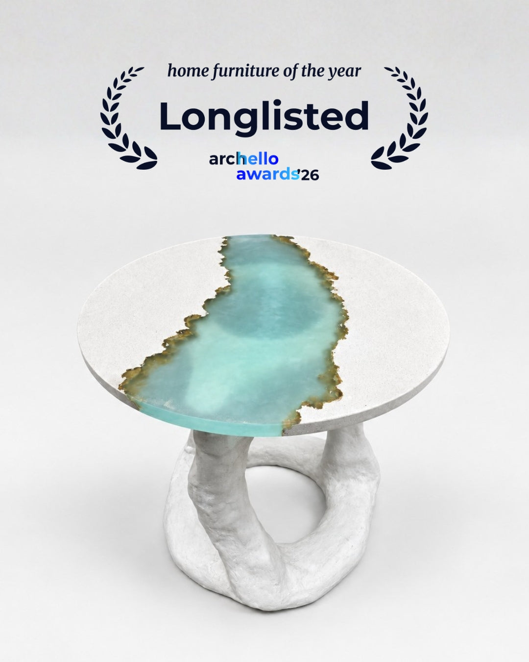 A round side table with a white stone-like concrete base and a turquoise blue resin inlay bordered in sand, shown on a light gray background with a laurel wreath and the text 'home furniture of the year Longlisted archello awards 26' above it.