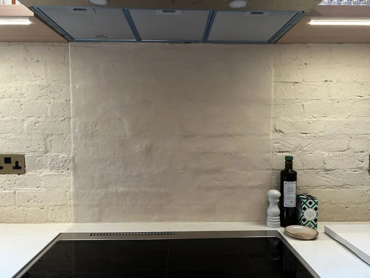 A textured beige microcemented splashback wall installed in a kitchen, featuring a black induction cooktop below and white brick walls on either side.