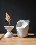 A modern sculptural white side table and matching curved lounge chair on a light wood floor against a dark charcoal wall, with a small vase of pampas grass on the table.