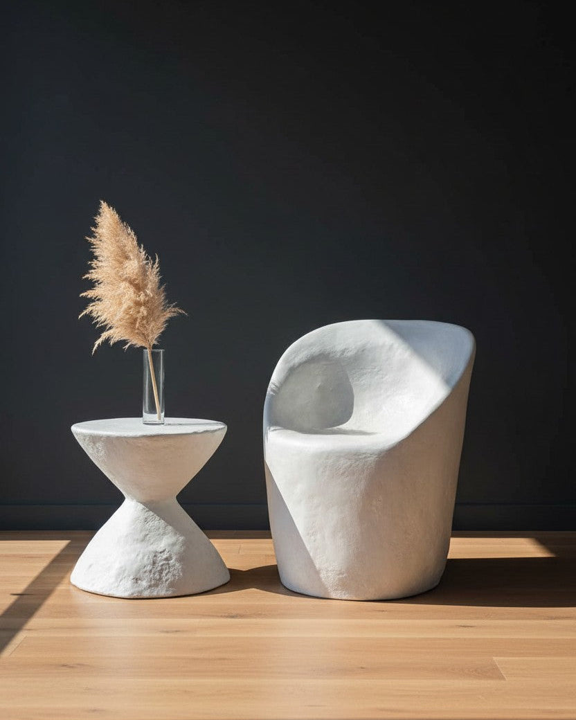 A modern sculptural white chair and matching side table with a vase of pampas grass, set on a light wood floor against a dark background.