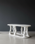 A sculptural white coffee table with a smooth oval top and organic, tree-like twisted legs, set against a dark gray background and light gray floor.