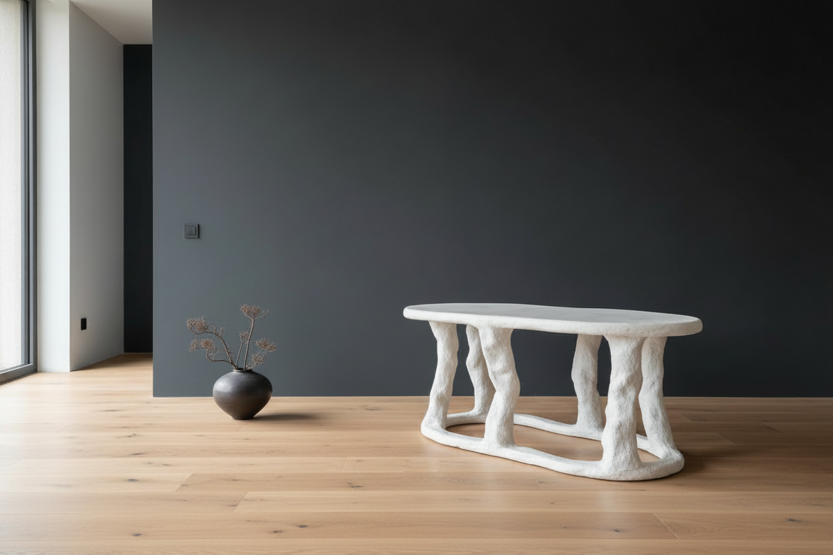 A modern organic-shaped white concrete table with sculptural, tree-like legs, set on a light wood floor against a dark charcoal wall. A small black vase with dried branches sits to the left.