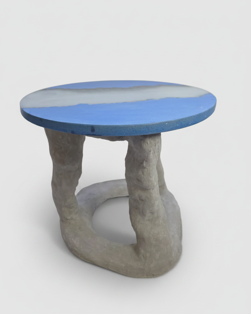A sculptural side concrete table with a round blue concrete and translucent resin top and a twisted concrete base, shown on a light gray background.