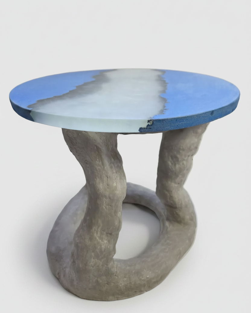 A sculptural round coffee table with a textured gray concrete base and a top featuring a blue and white wave-like pattern, shown on a light gray background.