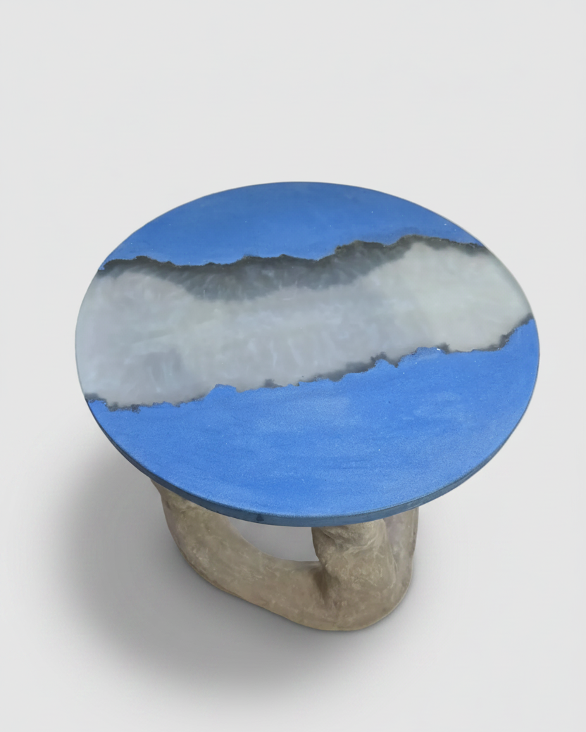 A round accent table featuring a vibrant blue top with a textured white and gray inlay, supported by a rustic, organic-shaped grey stone-like concrete base, shown on an off-white background.