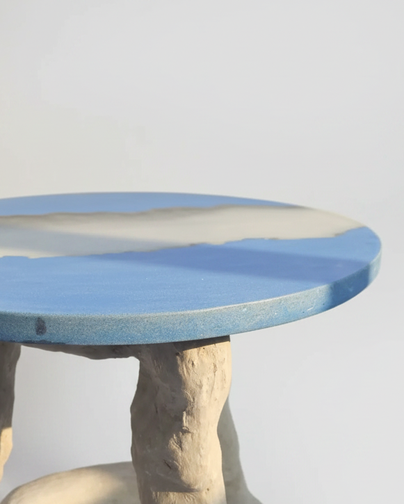 A close-up of a round table with a blue and beige resin-concrete top and textured light grey legs, photographed against a plain light gray background.